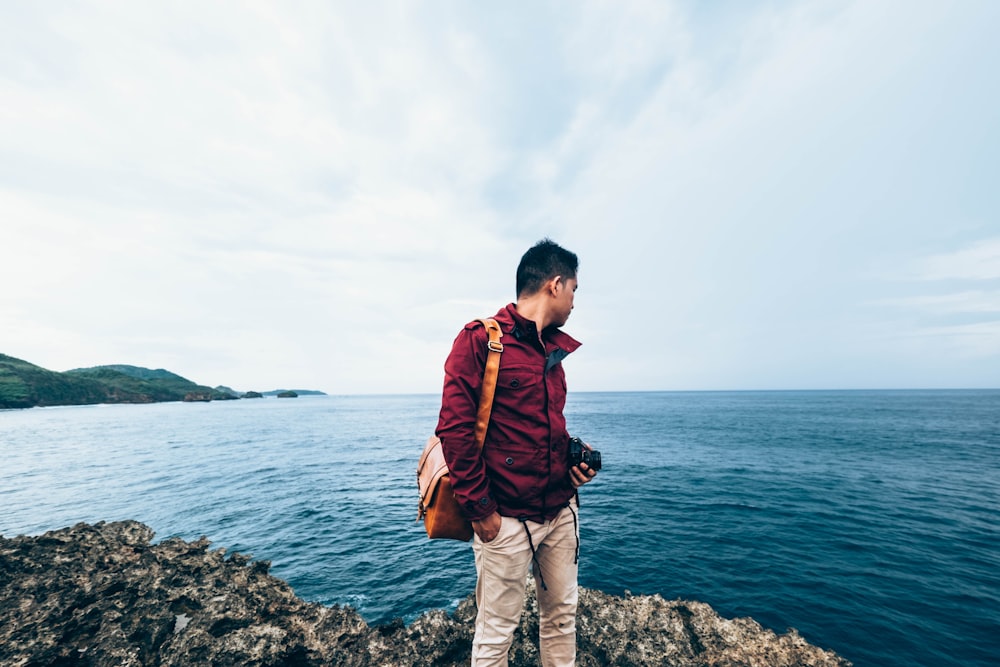 homem vestindo jaqueta de zíper vermelha em pé no topo do penhasco rochoso ao lado do corpo de água durante o dia
