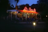 A vibrant outdoor cafe with a neon sign that reads 'La Boule' on its roof. There are several people gathered, some sitting and others standing, under the canopy. The atmosphere is illuminated by colorful string lights against the backdrop of a twilight sky.