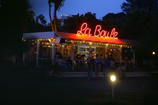 A vibrant outdoor cafe with a neon sign that reads 'La Boule' on its roof. There are several people gathered, some sitting and others standing, under the canopy. The atmosphere is illuminated by colorful string lights against the backdrop of a twilight sky.