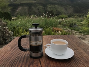 A rustic wooden table displaying a French press with freshly brewed coffee and a steaming cup beside it.