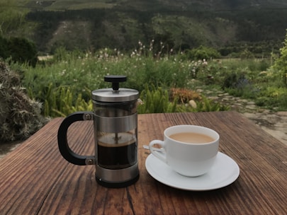 A rustic wooden table displaying a French press with freshly brewed coffee and a steaming cup beside it.