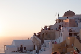 A picturesque setting with whitewashed buildings and a dome situated on a cliff overlooking the sea. The warm glow of the sunset casts a gentle light on the scene. Groups of people are gathered on the terraces, taking in the view. The architecture reflects a mixture of traditional Mediterranean and Cycladic styles, with narrow alleys and staircases.