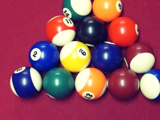 A cluster of vibrant billiard balls arranged in a triangular formation on a maroon surface. The balls display a variety of colors including blue, green, red, yellow, and orange, each marked with numbers.