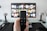 A person holds a remote control in front of a TV displaying a streaming platform interface with various movie and show thumbnails. The room includes a desk with two small lamps on either side of the TV.