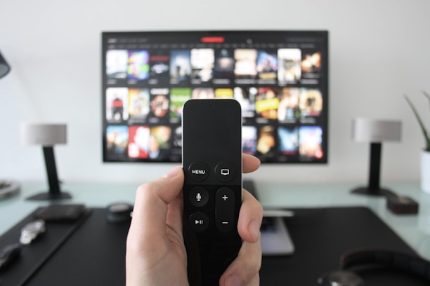 A person holds a remote control in front of a TV displaying a streaming platform interface with various movie and show thumbnails. The room includes a desk with two small lamps on either side of the TV.