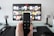 A person holds a remote control in front of a TV displaying a streaming platform interface with various movie and show thumbnails. The room includes a desk with two small lamps on either side of the TV.