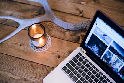 A wooden table features a lit candle in a glass holder next to a laptop displaying a webpage with images. An antler is partially visible, adding a rustic touch.