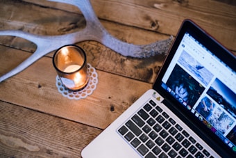 A wooden table features a lit candle in a glass holder next to a laptop displaying a webpage with images. An antler is partially visible, adding a rustic touch.
