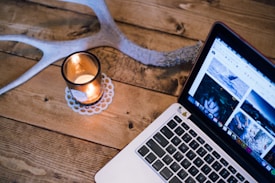 A wooden table features a lit candle in a glass holder next to a laptop displaying a webpage with images. An antler is partially visible, adding a rustic touch.
