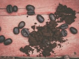 Coffee beans are scattered on a rustic wooden surface alongside a mound of ground coffee. The wood appears weathered and painted in a faded red color, contrasting with the dark, rich tone of the coffee.