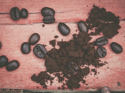 Coffee beans are scattered on a rustic wooden surface alongside a mound of ground coffee. The wood appears weathered and painted in a faded red color, contrasting with the dark, rich tone of the coffee.