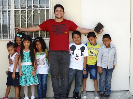 A group of children standing next to an adult man, who has his arms outstretched and is holding a book with his right hand. The children appear cheerful and the setting seems to be outside in front of a window with bars.