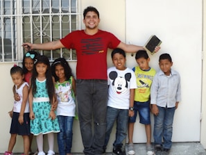A group of children standing next to an adult man, who has his arms outstretched and is holding a book with his right hand. The children appear cheerful and the setting seems to be outside in front of a window with bars.