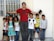 A group of children standing next to an adult man, who has his arms outstretched and is holding a book with his right hand. The children appear cheerful and the setting seems to be outside in front of a window with bars.