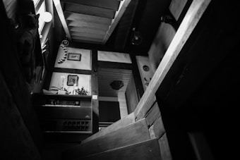 A black and white image of a wooden staircase leading to a landing. The setting is indoors, with a rustic and cozy interior design. Plants and wall decorations, including framed pictures and circular ornaments, adorn the space. Light filters in from a window on the left, creating a gentle contrast of shadows and highlights.