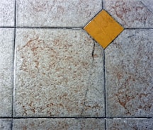 A textured floor surface with square tiles, featuring a distinct yellow diamond-shaped tile in the center. The surrounding tiles have a mottled light gray and brown appearance, with visible grout lines creating a grid pattern.