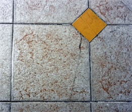 A textured floor surface with square tiles, featuring a distinct yellow diamond-shaped tile in the center. The surrounding tiles have a mottled light gray and brown appearance, with visible grout lines creating a grid pattern.