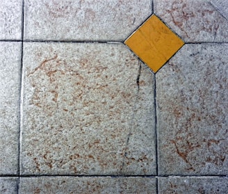 A textured floor surface with square tiles, featuring a distinct yellow diamond-shaped tile in the center. The surrounding tiles have a mottled light gray and brown appearance, with visible grout lines creating a grid pattern.