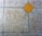 A textured floor surface with square tiles, featuring a distinct yellow diamond-shaped tile in the center. The surrounding tiles have a mottled light gray and brown appearance, with visible grout lines creating a grid pattern.