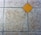 A textured floor surface with square tiles, featuring a distinct yellow diamond-shaped tile in the center. The surrounding tiles have a mottled light gray and brown appearance, with visible grout lines creating a grid pattern.