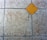 A textured floor surface with square tiles, featuring a distinct yellow diamond-shaped tile in the center. The surrounding tiles have a mottled light gray and brown appearance, with visible grout lines creating a grid pattern.