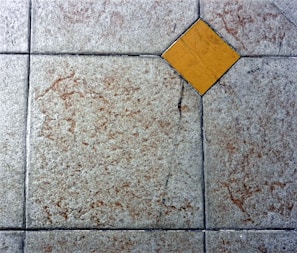 A textured floor surface with square tiles, featuring a distinct yellow diamond-shaped tile in the center. The surrounding tiles have a mottled light gray and brown appearance, with visible grout lines creating a grid pattern.