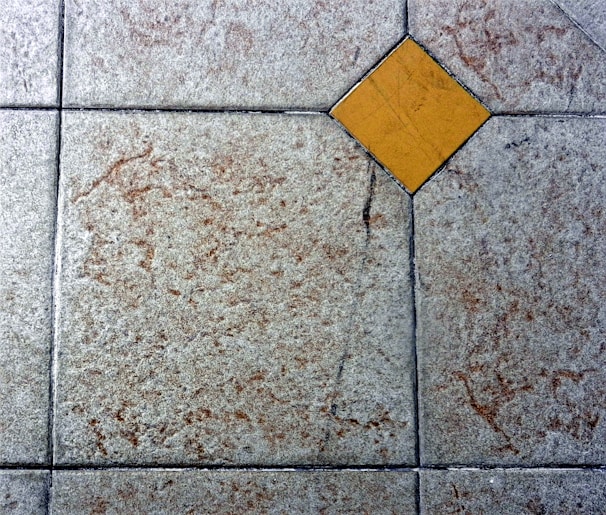 A textured floor surface with square tiles, featuring a distinct yellow diamond-shaped tile in the center. The surrounding tiles have a mottled light gray and brown appearance, with visible grout lines creating a grid pattern.