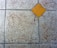 A textured floor surface with square tiles, featuring a distinct yellow diamond-shaped tile in the center. The surrounding tiles have a mottled light gray and brown appearance, with visible grout lines creating a grid pattern.