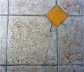 A textured floor surface with square tiles, featuring a distinct yellow diamond-shaped tile in the center. The surrounding tiles have a mottled light gray and brown appearance, with visible grout lines creating a grid pattern.