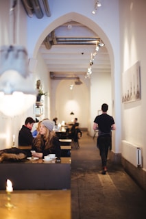 A warmly lit cafe interior with minimalist decor featuring arches, visible piping on the ceiling, and wall-mounted lighting. Patrons are seated at tables with coffee cups, including a woman with long hair and a knit hat looking off to the side and a man walking away down the aisle. Wall art and other decor elements contribute to the cozy atmosphere.