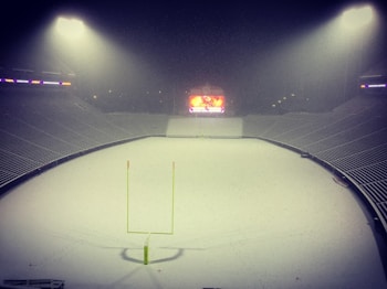 A football stadium covered in snow with a lit-up scoreboard at the far end. The floodlights illuminate the empty seats and the snow-covered field. A football goal post stands in the center of the field.