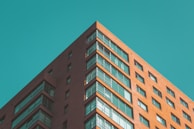 A modern high-rise apartment building with a reddish-brown brick facade and numerous windows reflecting the clear blue sky. The perspective is looking up from the corner of the building, giving prominence to its sharp angles and geometric lines.