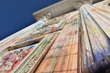 A tall classical building façade covered in colorful paint splatters and drips. The perspective is from below, emphasizing the height and ornate architectural details like columns and decorative carvings. The vivid paint colors contrast with the structure’s original white surface, creating an abstract, expressive appearance.