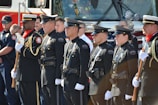 A group of uniformed police officers and other personnel stand in a line facing forward in a formal pose, suggesting a ceremonial or official gathering. They are dressed in dark uniforms with badges and other insignia, some holding ceremonial axes. There is a fire truck in the background, indicating the presence of firefighters as well.