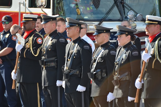 A group of uniformed police officers and other personnel stand in a line facing forward in a formal pose, suggesting a ceremonial or official gathering. They are dressed in dark uniforms with badges and other insignia, some holding ceremonial axes. There is a fire truck in the background, indicating the presence of firefighters as well.