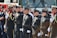 A group of uniformed police officers and other personnel stand in a line facing forward in a formal pose, suggesting a ceremonial or official gathering. They are dressed in dark uniforms with badges and other insignia, some holding ceremonial axes. There is a fire truck in the background, indicating the presence of firefighters as well.