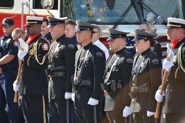 A group of uniformed police officers and other personnel stand in a line facing forward in a formal pose, suggesting a ceremonial or official gathering. They are dressed in dark uniforms with badges and other insignia, some holding ceremonial axes. There is a fire truck in the background, indicating the presence of firefighters as well.
