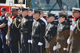 A group of uniformed police officers and other personnel stand in a line facing forward in a formal pose, suggesting a ceremonial or official gathering. They are dressed in dark uniforms with badges and other insignia, some holding ceremonial axes. There is a fire truck in the background, indicating the presence of firefighters as well.