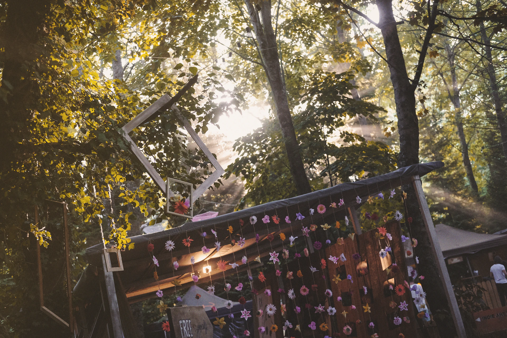 A serene outdoor gallery setup where framed mixed media pieces hang from tree branches, sunlight filtering through the canopy above.