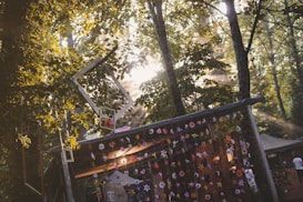 Sunlight filters through a dense canopy of trees casting dappled shadows across a rustic structure adorned with hanging flowers and frames. The atmosphere is serene and whimsical with an emphasis on natural beauty. Wooden textures and vibrant colors from the flowers add to the scene's charm.