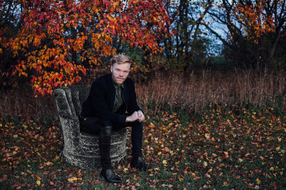 man wearing black suit sits on black concrete chair beside brown tree