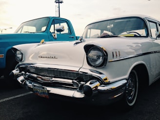 A classic white Chevrolet car parked next to a blue truck in an outdoor setting. The scene captures the vintage appeal with its chrome detailing and recognizable emblem on the grille. The vehicles are positioned diagonally, showcasing the front and side views.