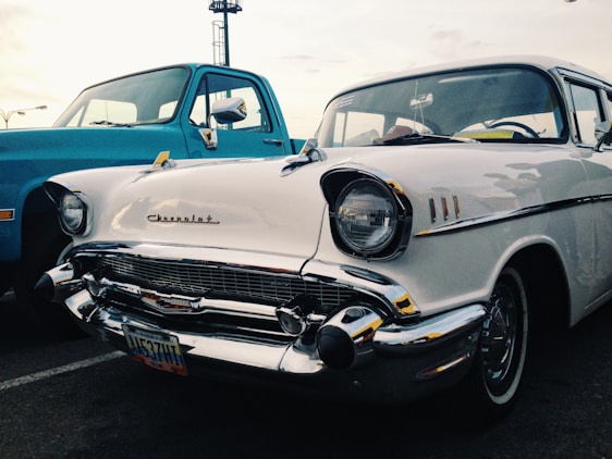 A classic white Chevrolet car parked next to a blue truck in an outdoor setting. The scene captures the vintage appeal with its chrome detailing and recognizable emblem on the grille. The vehicles are positioned diagonally, showcasing the front and side views.