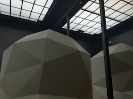 A room with a high ceiling featuring geometric, faceted structures in a light, neutral color. The ceiling consists of a grid of rectangular, translucent panels that allow light to filter through, creating a minimalist and modern aesthetic.