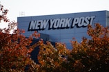 A building with the logo 'NEW YORK POST' prominently displayed on its exterior. In the foreground, autumn-colored leaves are visible under a clear sky.