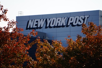 A building with the logo 'NEW YORK POST' prominently displayed on its exterior. In the foreground, autumn-colored leaves are visible under a clear sky.