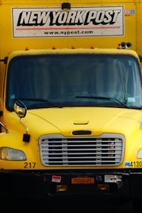 A yellow delivery truck with the 'New York Post' logo and website on the upper part of the vehicle. The truck has a prominent front grille, license plate, and various registration stickers.