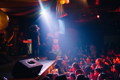A lively indoor scene with children and adults engaged in an event. The atmosphere is colorful and festive, with balloons and stage lighting. The crowd is large, with many people looking towards the stage where a person stands with their back to the camera. Musical instruments and stage equipment are visible.