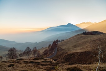 The image captures a serene and expansive landscape with rolling hills and a winding road that cuts through the terrain. In the background, distant mountains are softly illuminated by the early morning or late afternoon sun, creating a gentle glow. A layer of mist adds a dreamy quality to the scene, while sparse trees and brush dot the foreground, contributing to the peaceful atmosphere.