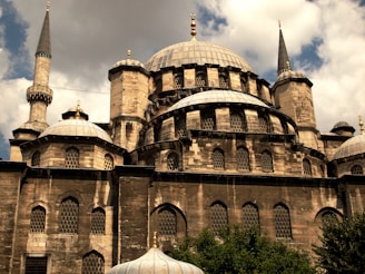 A historic mosque with multiple domes and tall minarets, constructed using stone and adorned with decorative elements. The architecture features intricate patterns on the window grilles and a blend of shadows and sunlight that highlights the texture of the stone. The sky is partly cloudy, adding depth to the scene.